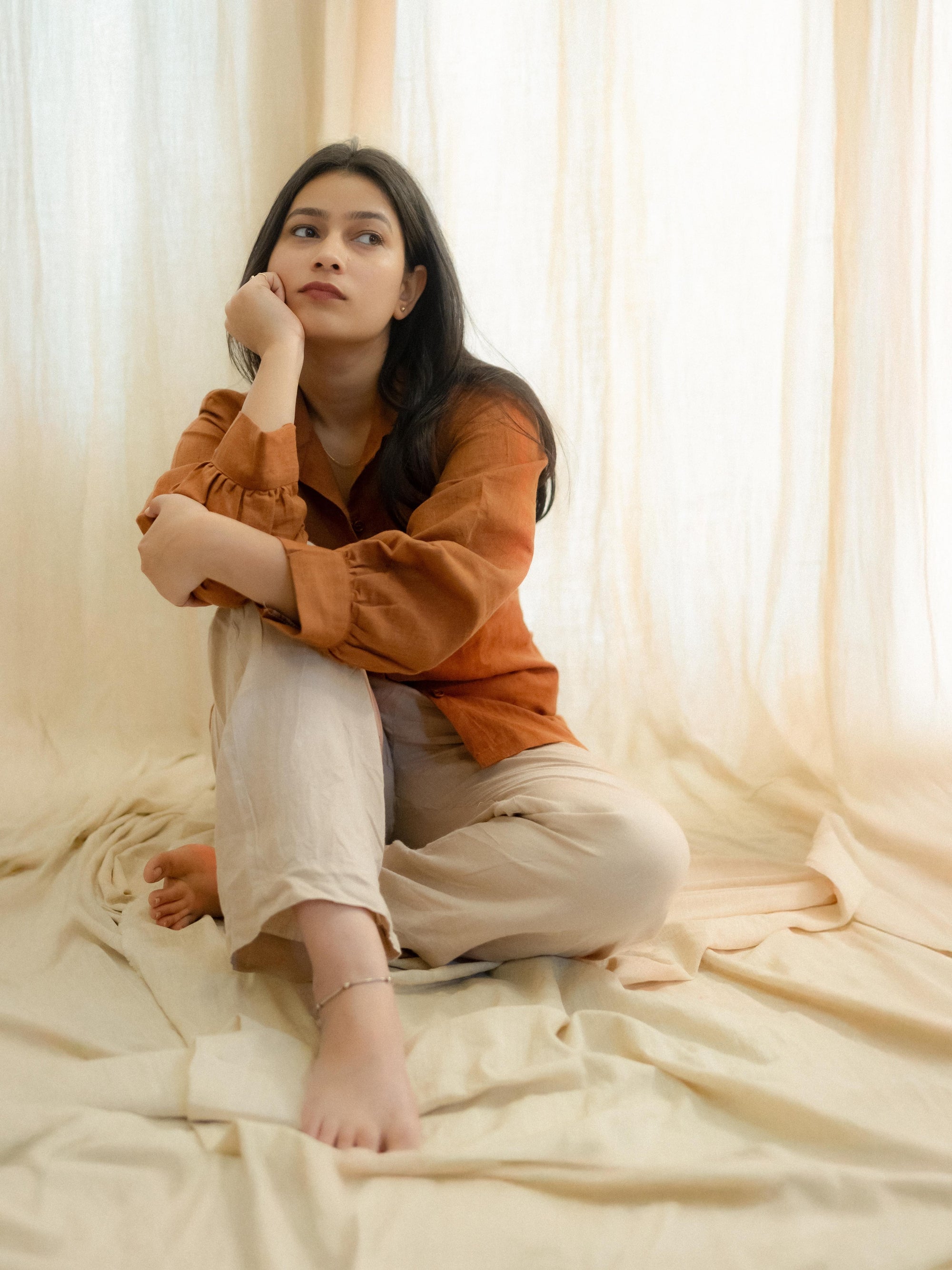 Woman sitting on a bed in a softly lit room with white curtains.